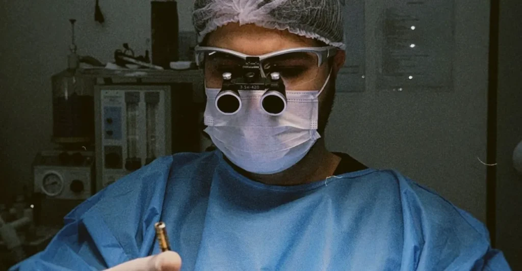 Dr. Éric Aguiar, médico, de touca, máscara e lupa cirúrgica em atendimento de transplante capilar